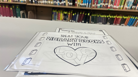 A stack of laminated signs lying on a white countertop. The top sign reads, in black letters, "Treat Your Library Books with Love." There are black outlines of books creating a border on the left and right side of the sign, and the word "Love" is inside the black outline of a heart. 