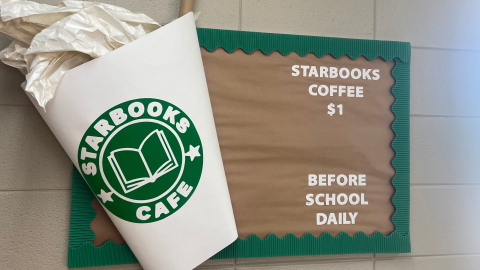 A bulletin board hangs on a beige tile wall. The bulletin board is decorated in brown paper with a green border, and features a large white coffee cup with a green logo in the style of Starbucks that reads, "Starbooks Cafe." The message on the bulletin board reads, "Starbooks Coffee $1 before school daily." 