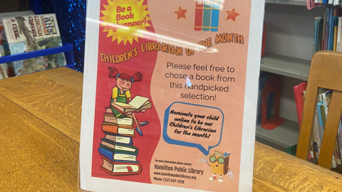 A standing acrylic frame holds a flyer advertising a contest for Children's Librarian of the Month. The sign reads, "Be a Book Influencer! Children's Librarian of the Month. Please feel free to chose a book from this handpicked selection! Nominate your child to be our Children's Librarian of the Month!" Contact information for the library is located at the bottom. 