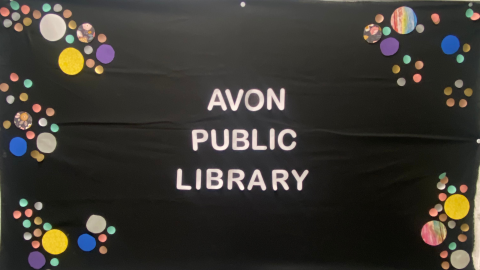 A bulletin board covered in black paper with circles of various sizes and colors along the right and left edges. In the center, it reads, "Avon Public Library" in white letters. 