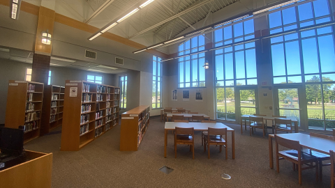 The library space featuring several sets of wooden tables and chairs in front of floor-to-ceiling windows. To the left of the tables and chairs are three rows of bookshelves holding books. 