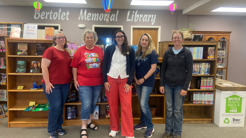 Five people standing in front of bookshelves holding library books and other materials. Behind them is a grey wall with the words, "Bertolet Memorial Library" painted in large, white letters. Below the words are two doors, the door on the left is open. To the right and left of the doors, wooden display cases sit against the wall. 