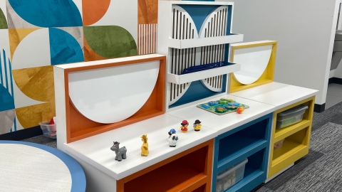 A childrens play area decorated in orange, blue, and yellow featuers white countertops and shelves and bins of children's toys. 