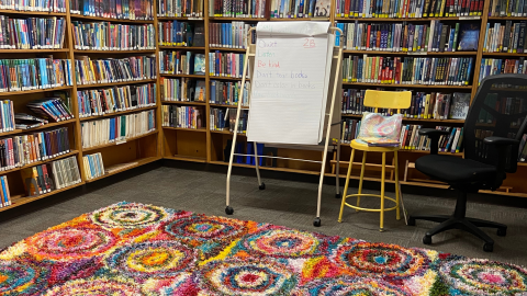 The library's reading corner, with shelves of books lining the walls, and a colorful, geometric rug on the floor. A rolling stand holds a pad of flipchart paper, and the top page reads, "Quiet. Listen. Be kind. Don't tear books. Don't color in books, Use bookboards. 2B." A yellow stool and black office chair are to the right of the flipchart. 