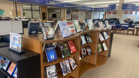 The Carl Sandburg College Library containing low display bookshelves with movies books on easels, a tech help area, tables and chairs for students, and a circulation desk off to the left. 