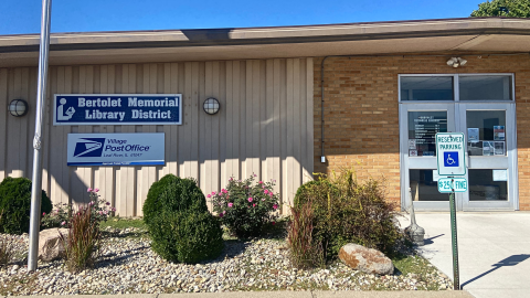 The facade of the building housing the Bertolet Memorial Library District and the Village Post Office for Leaf River, Illinois. The left side of the building is beige metal siding, and the right features a light brown bricks.  A sidewalk leads to the double, glass door entrance on the right, and a landscape bed containing rocks, various plants, and a flag pole is located in front of the building on the left. 