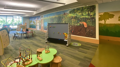 An indoor children's play area featuring wooden play stations and several tables with computer stations. The far wall features a large panoramic image of a prairie grassland with plants and animals above and below ground. 