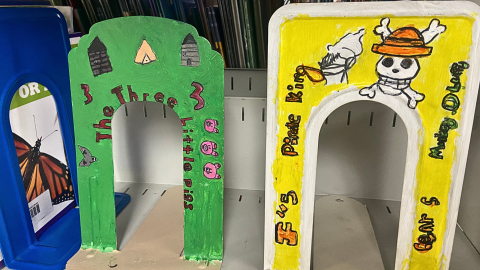 Three painted metal bookends sit on a metal shelf. The bookend on the far left is blue and sits under a library book. The other two book ends face out. The bookend on the left is painted green and has images from the story of The Three Little Pigs. The bookend on the right is painted bright yellow with a white border and includes orange, black, and white imagery and words for a pirate story.