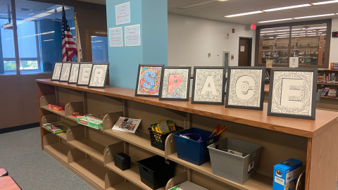 A row of low wooden bookshelves sits in front of a light blue pillar. On top of the shelves are 10 black frames containing a single capital letter surrounded by doodle art that spell out the words, "Maker Space." The shelves hold bins with various games, crafts, and maker space items. 