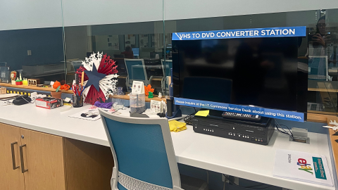 The library's VHS to DVD Converter station consisting of a monitor and VHS/DVD player sits on top of a white table top. In front of the table is a teal blue office chair, and to the left of the monitor are various office supplies and display items. 