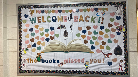 A bulletin board display featuring on open book at the bottom with various colored hearts rising up from it. Among the hearts are cutouts of various children's book characters. The top of the bulletin board reads, "Welcome Back!", while a message at the bottom says, "The books missed you!"