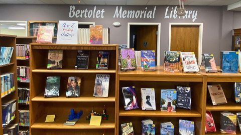 Wooden bookshelves holding a variety of books displayed on easels. Behind the bookshelves is a grey wall with the words, "Bertolet Memorial Library" in large, white letters. Beneath the words are two doors, and the door on the left is open. 