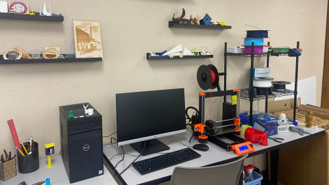 The library makerspace includes two gray tables holding a cup of pens, a desktop computer and monitor, a 3D printer, and a small, black, wire rack that contains spools of plastic thread in various colors. On the beige wall above the equipment are four black display ledges holding a variety of plastic and wood makerspace projects.