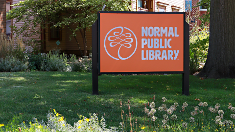 An outdoor orange sign that reads Normal Public Library is surrounded by wildflowers. 