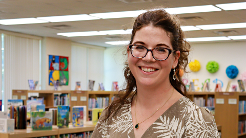 Jessica Sparenberg stands in the center of the library, smiling at the camera. 