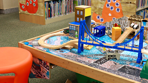 A wooden toy train set on a low table in front of colorful bookshelves.  