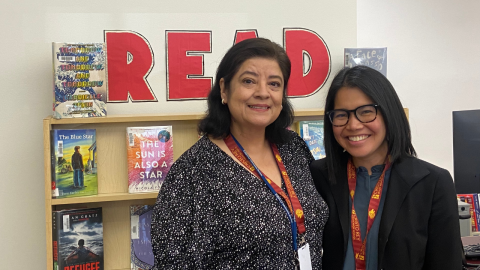 Two individuals standing in front of a low, wooden bookshelf holding library books. Two books are displayed on the top shelf, and between these books red capital letters spell out the word "Read."