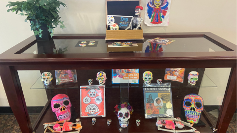 A short, glass display case holding a Day of the Dead display. The shelves in the case hold skulls of varying sizes that have been painted and decorated with flowers, as well as books about the holiday. On top of the display case is a box holding a sitting skeleton, and a sign that reads, "Day of the Dead." To the left of the box is a black vase holding a plant. On the wall above the case, a painted artwork of an angel hangs on the wall. 