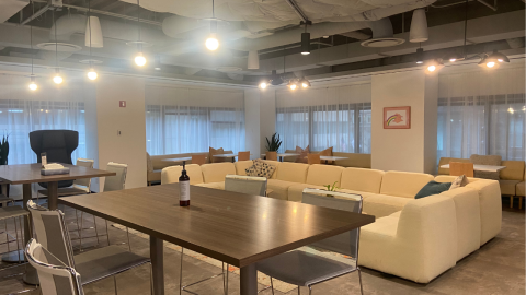 A common area featuring a yellow, u-shaped sectional sofa behind two high-top wooden tables with stools. Behind the sofa, tables and chairs line the wall full of windows. Sheer, white curtains hang over the windows. 