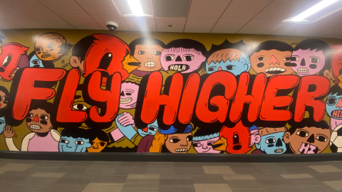 A graffiti-style mural featuring illustrated, cartoon-style heads representing a diverse population. Written in bubble style red letters with a black shadow, it reads, " Fly Higher."