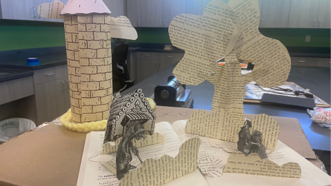 A tabletop display depicting a scene from Hansel & Gretel which features a paper tower, a cottage, and characters. Clouds, a tree, and bushes are cut from book pages. 