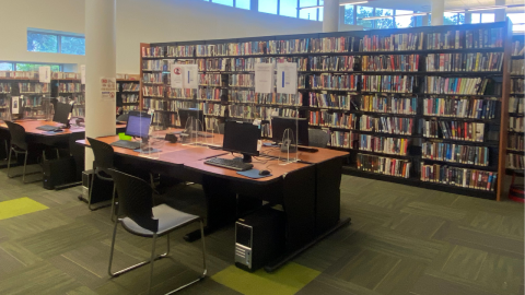 A view of the library featuring tall, freestanding, wooden bookshelves holding library books. In front of the shelves are computer workstations with chairs and instructions for use. Beyond the shelves are more bookselves and windows. 