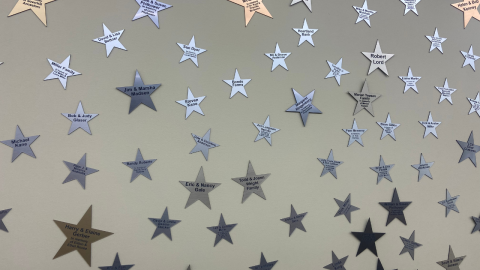 Multi-size metal stars hanging on a cream colored wall. In black print text, each star bears the name of supporters of the library, both individuals and organizations. 