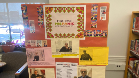 A bulletin board covered in dark orange paper features a National Hispanic Heritage Month display. In the center is a sign reading, "National Hispanic Heritage Month," and on either side of the sign are images of prominent Hispanic individuals. The lower half of the display features papers sharing bios of famous Hispanic figures. 