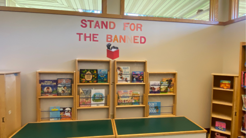 A banned books display in the library features two shallow, wooden bookshelf holding banned children's books. Above the display, in red capital letters, are the words, "Stand for the Banned," along with a cutout of an individual with a raised fist, reading a book with a red cover.  