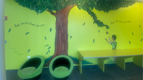 A mural of a scene from The Giving Tree features an apple falling into the outstretched hands of a boy. On the left side of the tree trunk it reads, "And the boy loved the tree.."; on the right are the words, "And the tree was happy." In front of the wall are two, short, round green chairs and a yellow bench. 