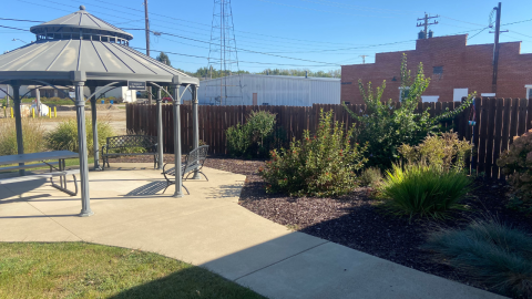 A courtyard area near the library featuers a metal gazebo with a table and benches underneath. A concrete walkway lined with bushes and shrubs leads to the gazebo. Beyond the gazebo is a brown picket fence; nearby brick and metal buildings and power lines are visible behind the fence. 