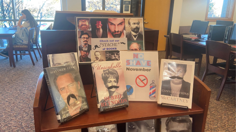 A no-shave November book display sits on top of a wooden bookshelf. A sign featuring images of people with mustaches reads, "Check out our "STACHE" of books." Four memoirs featuring covers with headshots sit in display easels with a black paper mustache added to the person on each cover.  