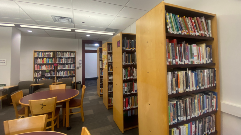 A view of the library with four, tall wooden bookshelves full of library books situated perpendicularly along the right wall. To the left of the bookshelves are tables with chairs, and along the far wall is another bookshelf on the left and a door on the right. 