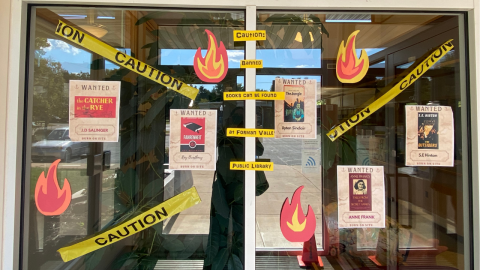 Glass double, sliding doors to the library featuring a banned books display. Caution tape extends from corner to corner, with the words, "Caution: Banned Books Can Be Found at Forman Valley Public Library" in black on yellow paper. Wanted signs for banned books hang among flames made of red, orange, and yellow construction paper. 