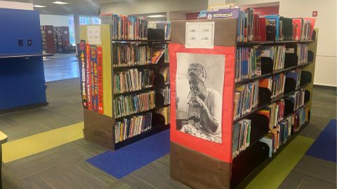 Two rows of short wooden bookshelves holding library books. On the end of the shelf on the left are oversized, paper representations of book spines of The Hunger Games, The Adventures of Captain Underpants, Harry Potter and the Sorcerer's Stone, and The Hate U Give. On the end of the shelf on the right is a photo poster of a woman holding the book "I Know Why the Caged Bird Sings." Both shelves feature wayfinding signs. 