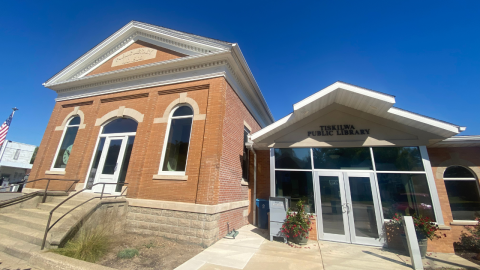 The front of the Tiskilwa Public Library, an orange-brown brick building with a white roof and trim. The glass, double-door entrance to the library is on the right; a concrete sidewalk leads up to the doors. 