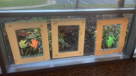 Three light brown, paper frames hang in the lower portion of a library window. In the first frame are two handprints, the left yellow and the right orange, a blue stick figure, and green grass drawn in window markers. Above the hands in red are the words, "happy Thanksgiving." The frame all the way to the right features a green pine tree with a brown trunk and red dots drawn with window markers. 