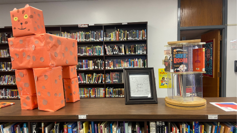 A short, wooden library bookshelf full of books; on top of the bookshelf is an orange tiger made of boxes covered in orange paper, a framed illustration reading "Spooky Books! Horror" with outline drawings of ghosts, and a plastic spinner of  books. 