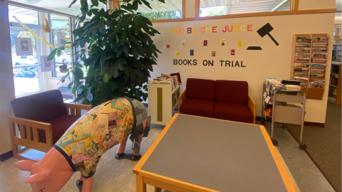 A banned books display inside the library entrance featuring images of banned books hanging under the words, "You Be the Judge, Books on Trial." Below the words is a red two-seat sofa with a book cart full of books on either side. To the right of the words, a magazine rack hangs on the wall. In front of the sofa, is a table, and a large pig sculpture covered in decoupage. To the left of the words is a fake tree. 