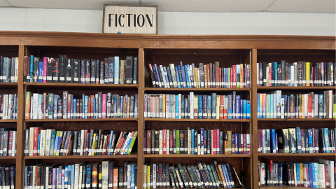 A wall of tall, wooden bookshelves holding library books. Above the bookshelves is a framed sign reading, "Fiction" in black letters on a white background. 