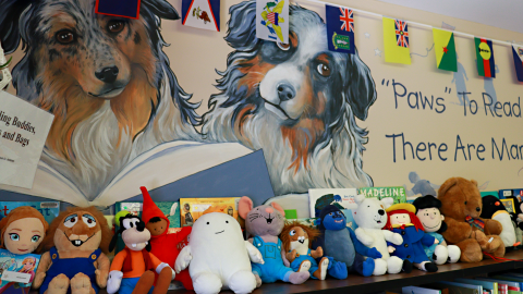 A mural of a dog holding a book. A row of stuffed animals on top of low bookshelves. 