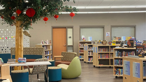 The seating area of a middle school library with benches, tables, and chairs surrounding a fake tree in the center. To the right of the seating area are shelves of low bookshelves full of library books with some displayed in easels on top. 