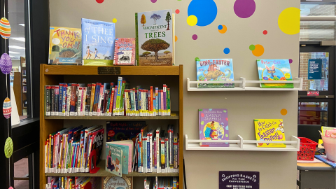 A library bookshelf display of children's books in front of a tan wall decorated with multi-colored, multi-sized circles