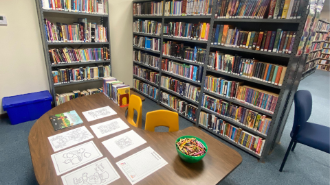 A u-shaped table and chairs for children holds coloring pages and a bowl of crayons. Behind the table are tall library bookshelves full of books. 