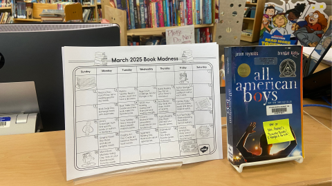 A paper calendar displays the activities for the March 2025 Book Madness. The book All American Boys by Jason Reynolds & Brendan Kiely sits in an easel to the right. 