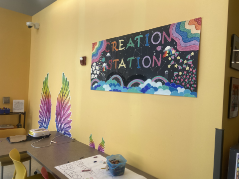 The library's creation station features sunny yellow walls, wood tables and chairs. A coloring work in progress is visible on the table.  
