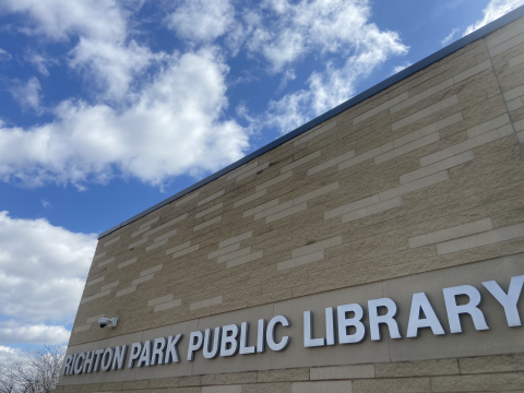 Richton Park Public Library sign hangs on a tan brick building visible under a blue, cloudy sky