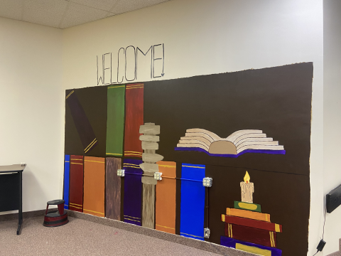 A welcome mural painted on an off-white shows book spines as if displayed on a bookshelf. The right side of the mural features an open book and a stack of books with a candle on top. 