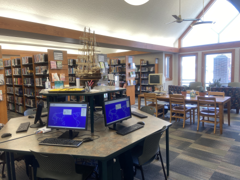 A patron computer workstation featuring a hexagon table, chairs, monitors, and keyboards. Beyond the workstation, library shelves full of books are visible. 