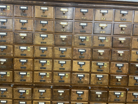 A vintage, wooden, library card catalog featuring rows of numberd and labeled drawers with metal handles. 